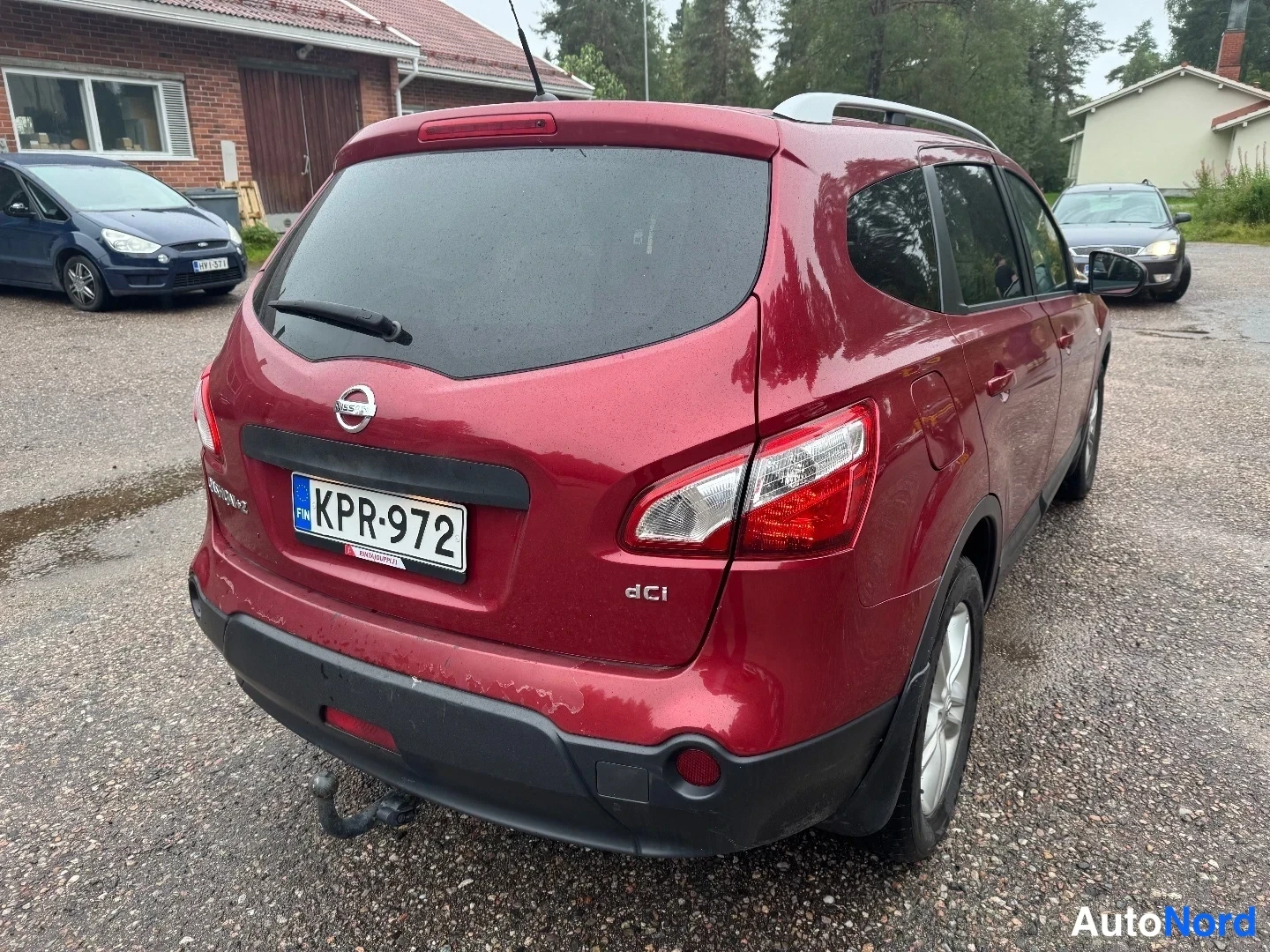 Takaviisto: Punaisen Nissan Qashqai+2:n takakulmanäkymä, rekisteritunnus, kattokaiteet ja vetokoukku näkyvissä.