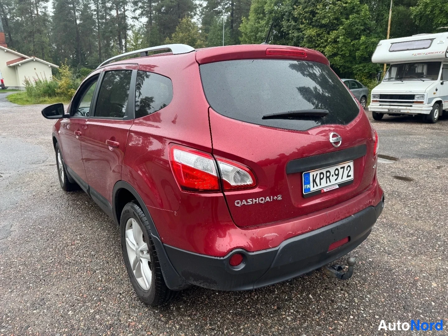Takaviisto: Punainen Nissan Qashqai+2 kuvattuna takaa ja sivulta: takaosassa Nissan-merkki, QASHQAI+2-teksti ja rekisterikilpi, katolla kattokaiteet ja takapuskurin alla ve