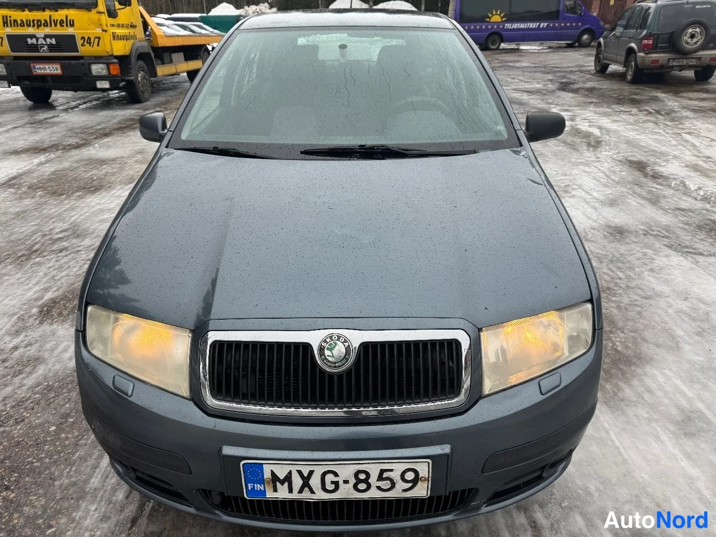 Edestä: Sinertävän harmaa henkilöauto kuvattuna suoraan edestä: etusäleikkö ja merkki, molemmat ajovalot sekä rekisterikilpi näkyvät. Tuulilasi ja sivupeilit ovat näkyv