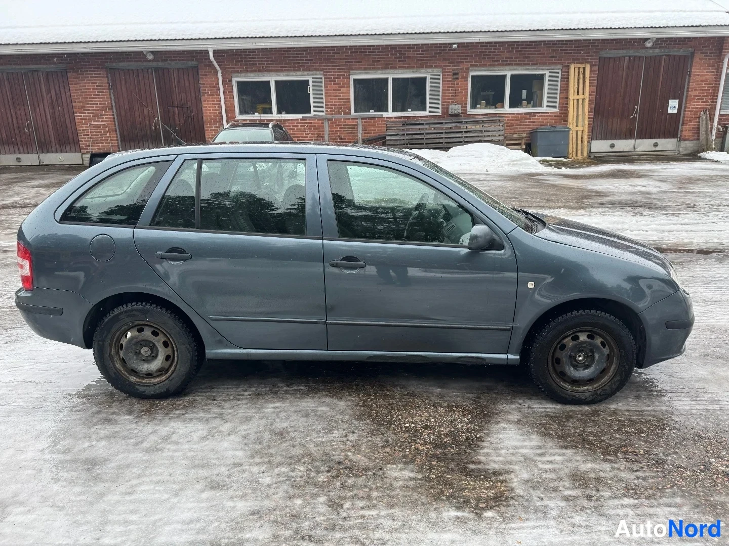 Takaviisto: Harmaansininen farmariauto sivulta takaa kuvattuna lumisella pihalla, tiilirakennus taustalla; laadukas mutta matalaresoluutioinen kuva.