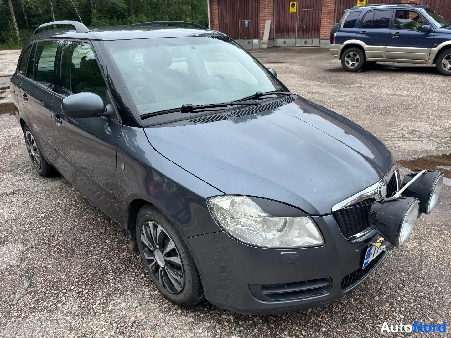 Etuviisto, nokka oikealle: Tummanharmaa farmariauto etuviistosta, keula kuvan oikeassa laidassa; keulassa kaksi pyöreää lisävaloa ja katolla kattokaiteet.