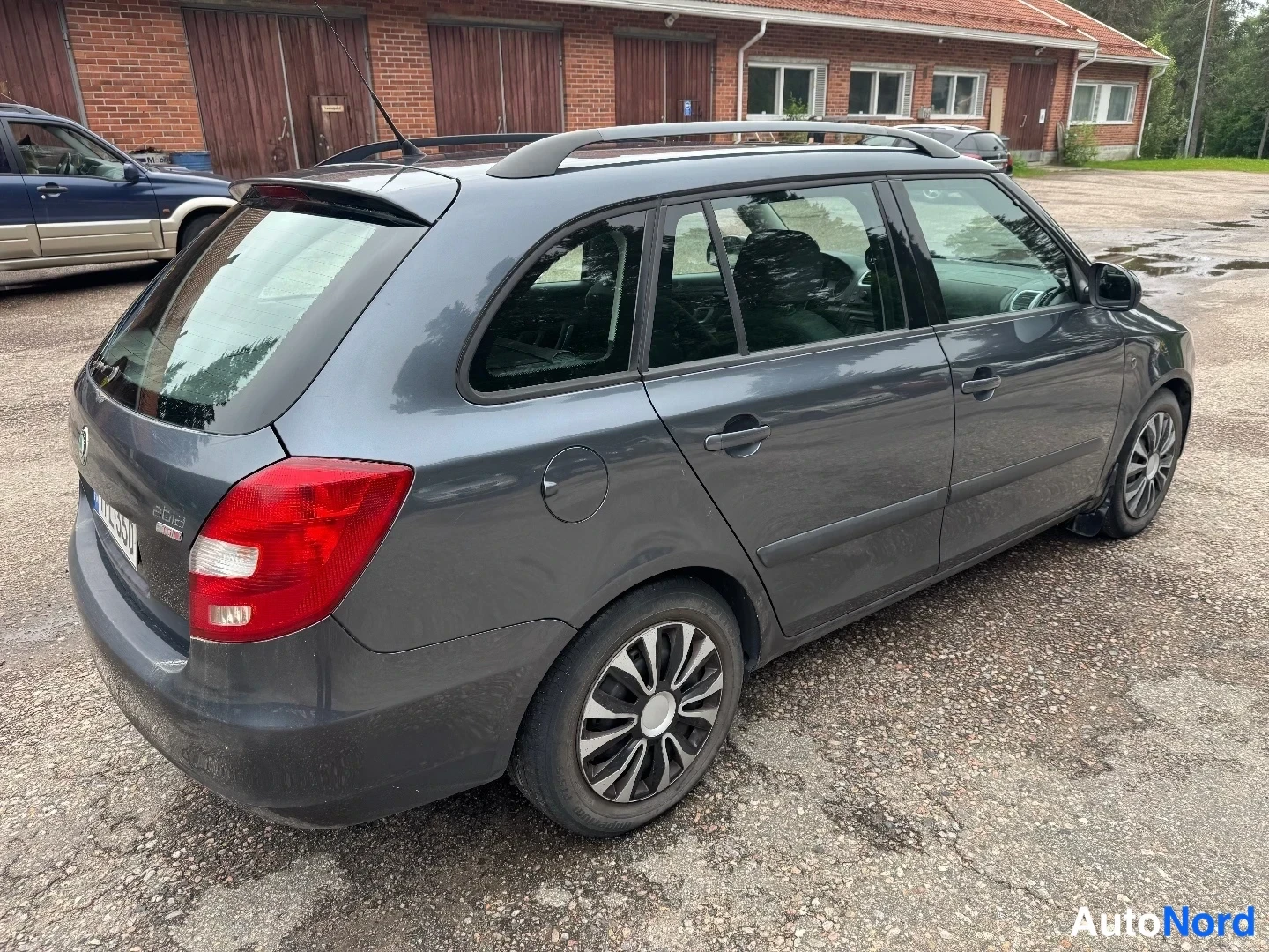 Takaviisto: Tummanharmaan farmariauton takakulmanäyttö: punaiset takavalot, kattokaiteet ja kevytmetallivanteet näkyvät, auto on pysäköity sorapihalla.