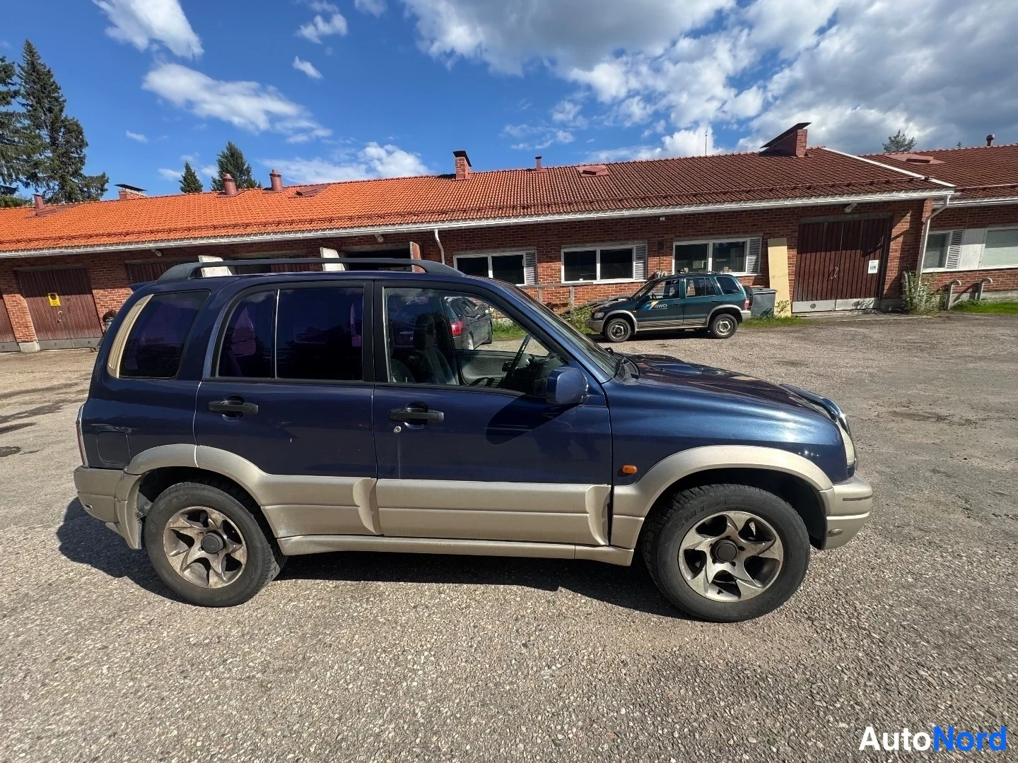 Etuviisto, nokka oikealle: Tummansininen Suzuki Grand Vitara etukolmannelta, nokka oikealla; vaaleanbeige aluslista, kattokaiteet ja tummat lasit; taustalla punatiilinen matala rak