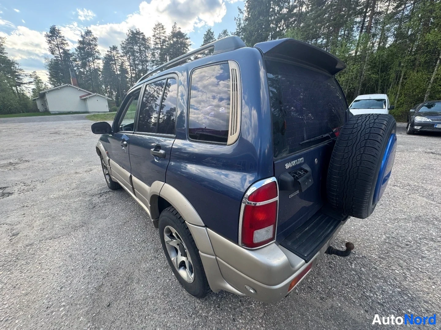 Takaviisto: Tummansininen Suzuki takaa ja vasemmalta sivulta viistosti kuvattuna: varapyörä takaluukun kannessa, kattokaiteet, kattospoileri, takavalot ja rekaviisi näkyvät