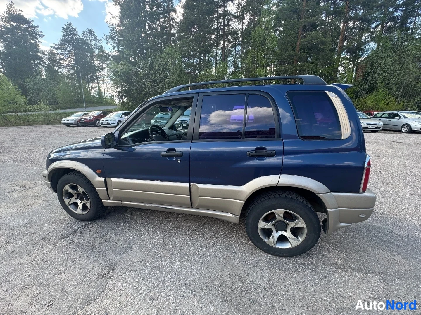 Takaviisto: Sisäinen kuva tumman sinisestä SUV-autosta nähtynä takaa ja oikealta kulmassa. Auto on pysäköity asfaltilla metsän reunassa. Takaluukku ja taka- sekä sivuvalot