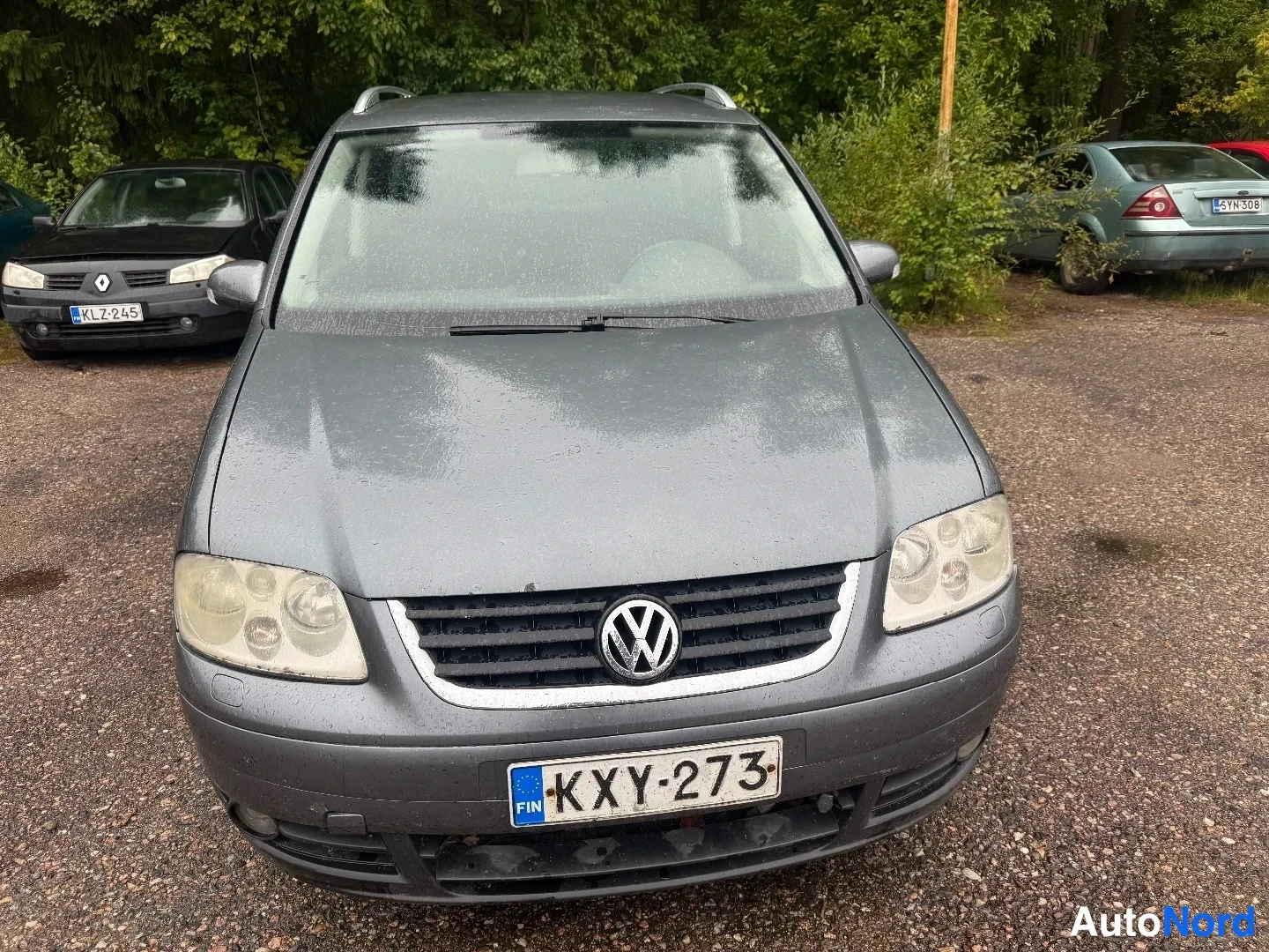 Edestä: Suora etunäkymä harmaasta Volkswagen Touranista; rekisterikilpi lukee, kattokaiteet ja sumuvalot näkyvät, kori on sateen kastama.