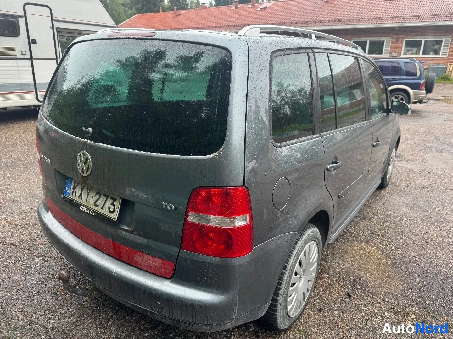 Takaviisto: Tummanharmaan Volkswagen-henkilöauton takaosa ja oikea kylki vinosti takaa kuvattuna. Takaluukussa VW-merkki ja rekisterikilpi. Katossa kattokaiteet ja takaalai