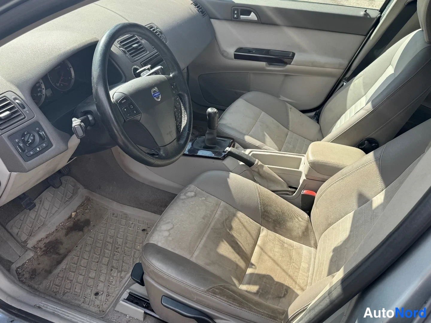 Ohjaamo: Beige front seats and dashboard area viewed from the open passenger door; steering wheel has controls, manual gear lever, and handbrake present. The driver-side