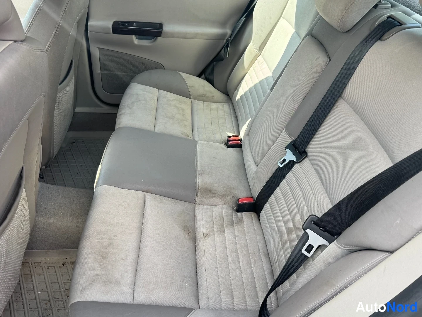 Takaistuimet: Close-up of the beige cloth rear bench seats in a car interior, showing dirt marks on the seat base and seatbelt buckles visible against the fabric.