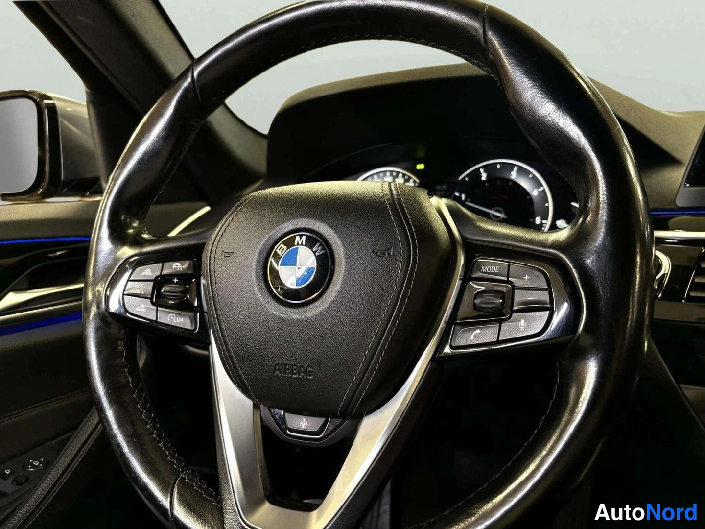 Muu: BMW-ohjauspyörä lähikuvassa, keskellä BMW-embleemi ja AIRBAG-teksti, vasemmalla LIM- ja rattempti- ja oikealla MODE-, puhelin- ja äänikomennot; taustalla tacho-