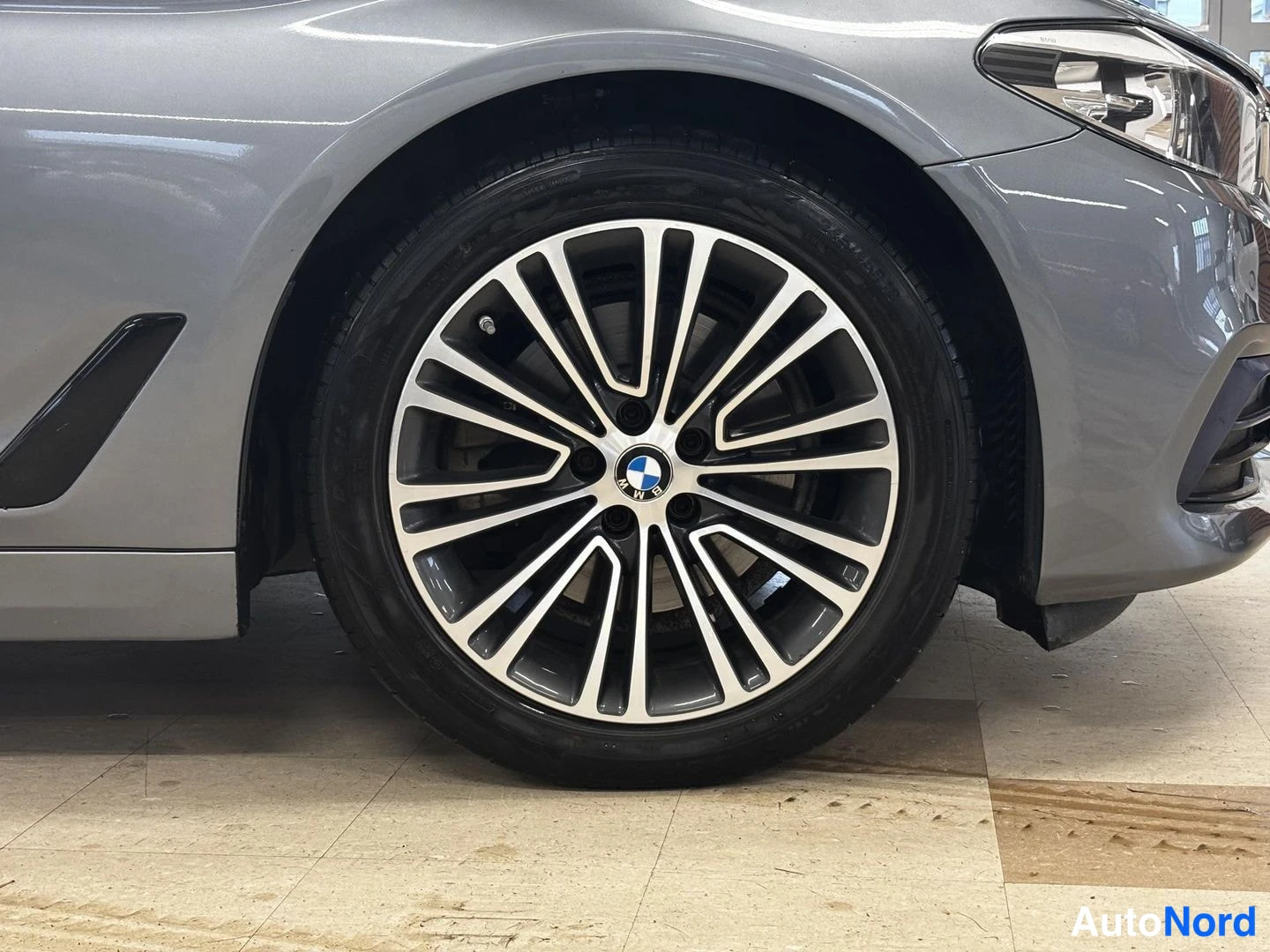 Etuviisto, nokka oikealle: Harmaan auton etupyörän kevytmetallivanne monivarsimallilla ja BMW-merkki nappikeskiössä; ajovalo näkyy kuvan yläreunassa.