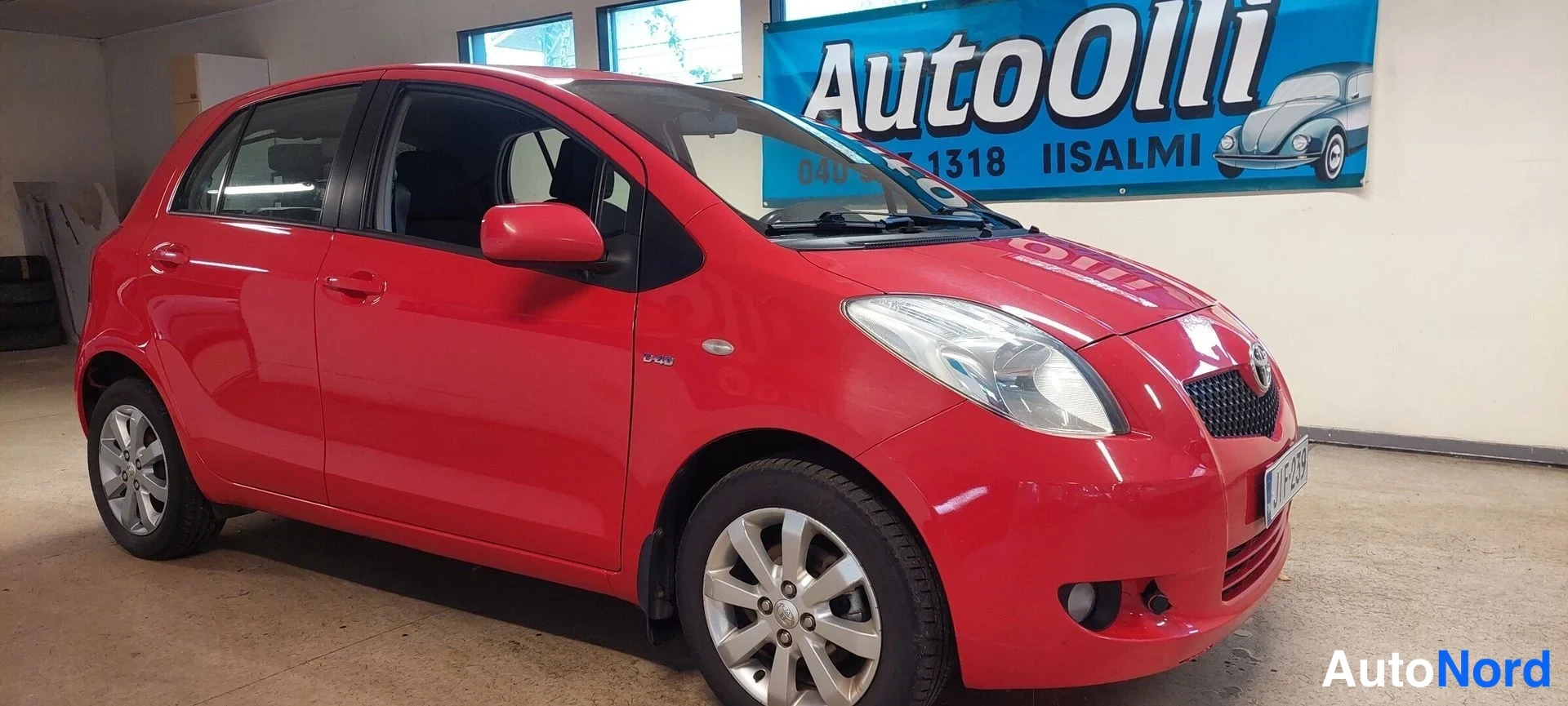 Etuviisto, nokka oikealle: Punainen Toyota Yaris -henkilöauto etuviistosta oikealta, takana AutoOlli-mainoskyltti.