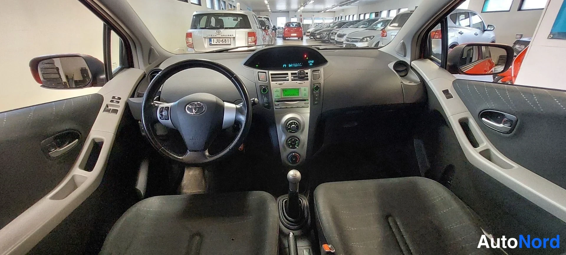 Etuistuimet: Etusisäkuvaa Toyota Yaris -mallin ohjaamosta. Ohjauspyörässä on Toyotan logo, keskellä mittariyksiköllä on vihreä taustavalo. Etupenkit ovat verhoiltuna mustall