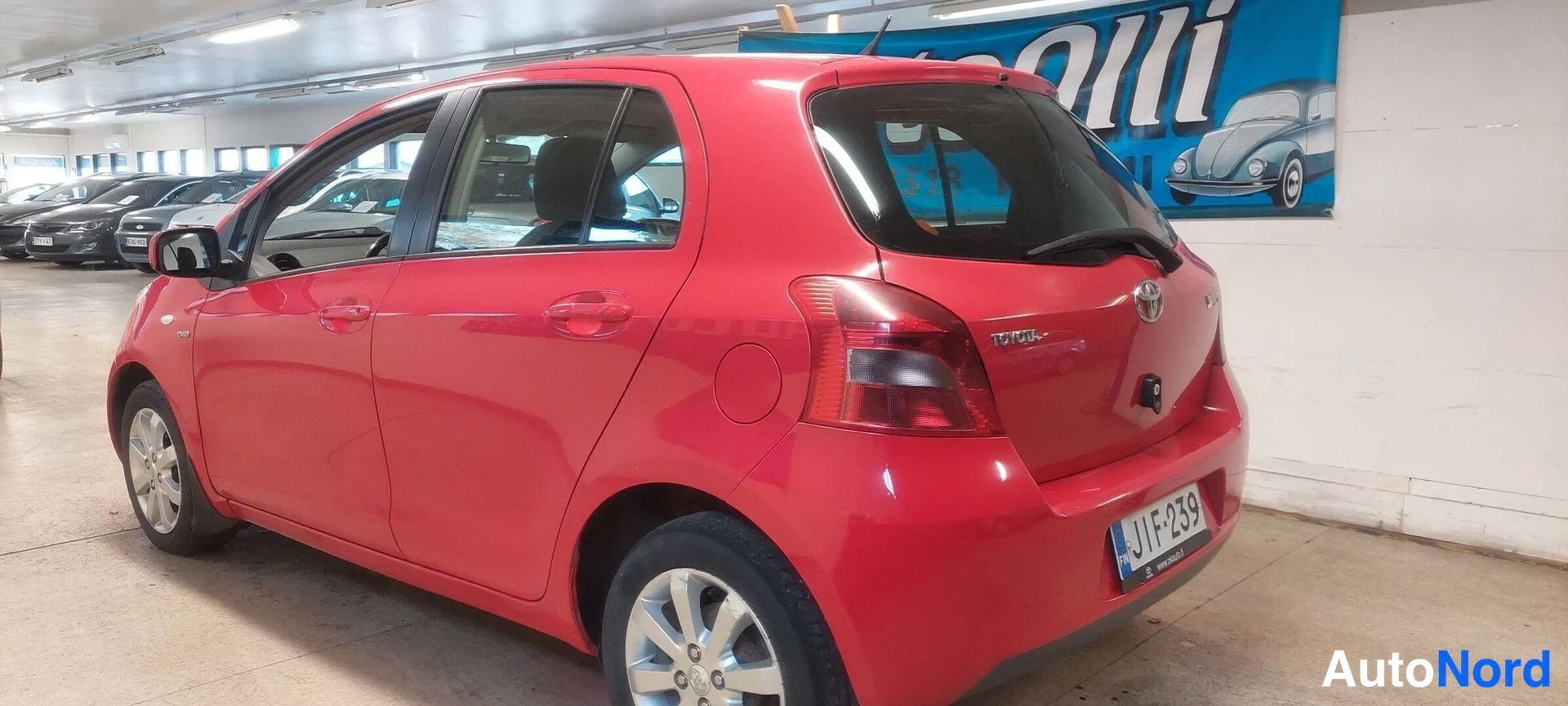 Takaviisto: Punainen Toyota Yaris -henkilöauto takaviistosta vasemmalta, rekisterikilpi näkyvissä.