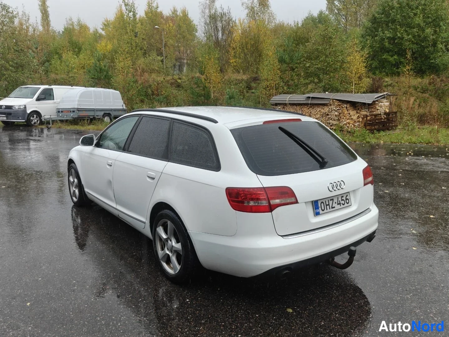 Takaviisto: Valkoinen Audi A6 Avant -farmari takavartinäkymässä vasemmalle päin, rekisteritunnus, märällä pihalla.