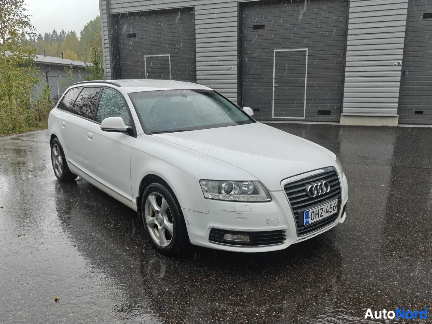 Etuviisto, nokka oikealle: Valkoinen Audi A6 Avant -farmari kolmivartinäkymässä keula oikealla, rekisteritunnus, sateella autotallin edustalla.