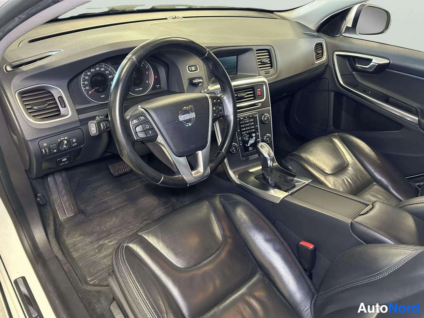 Muu: Volvo V60:n ohjaamo kuvattuna kuljettajan oven suunnasta: ohjauspyörä, mittaristo, keskikonsoli ja mustat nahkaistuimet.