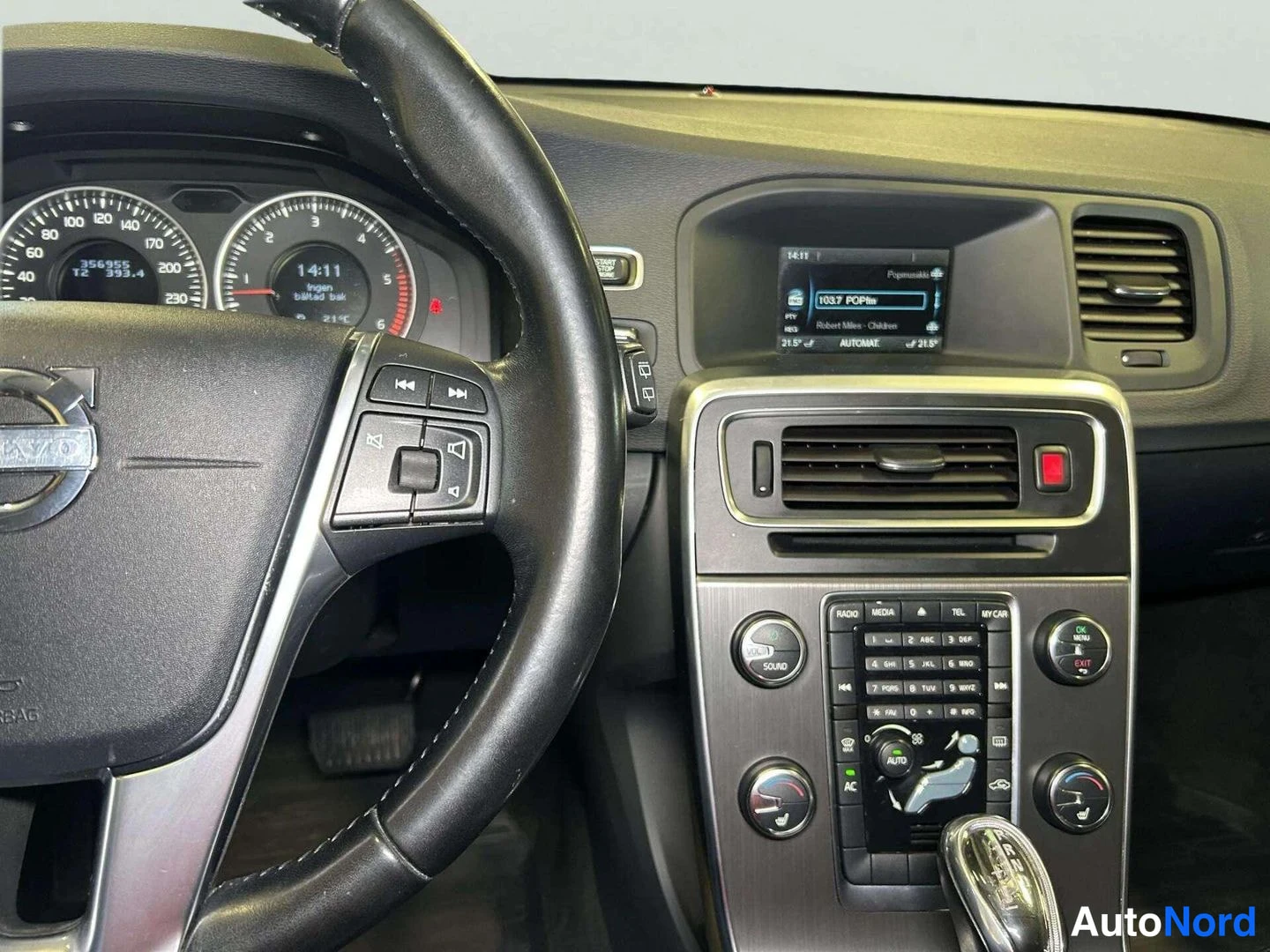 Muu: Ohjaamon näkymä: Volvo-ohjauspyörä, mittaristo ja keskikonsoli, jossa radiopaneeli ja automaattivaihteen valitsin.