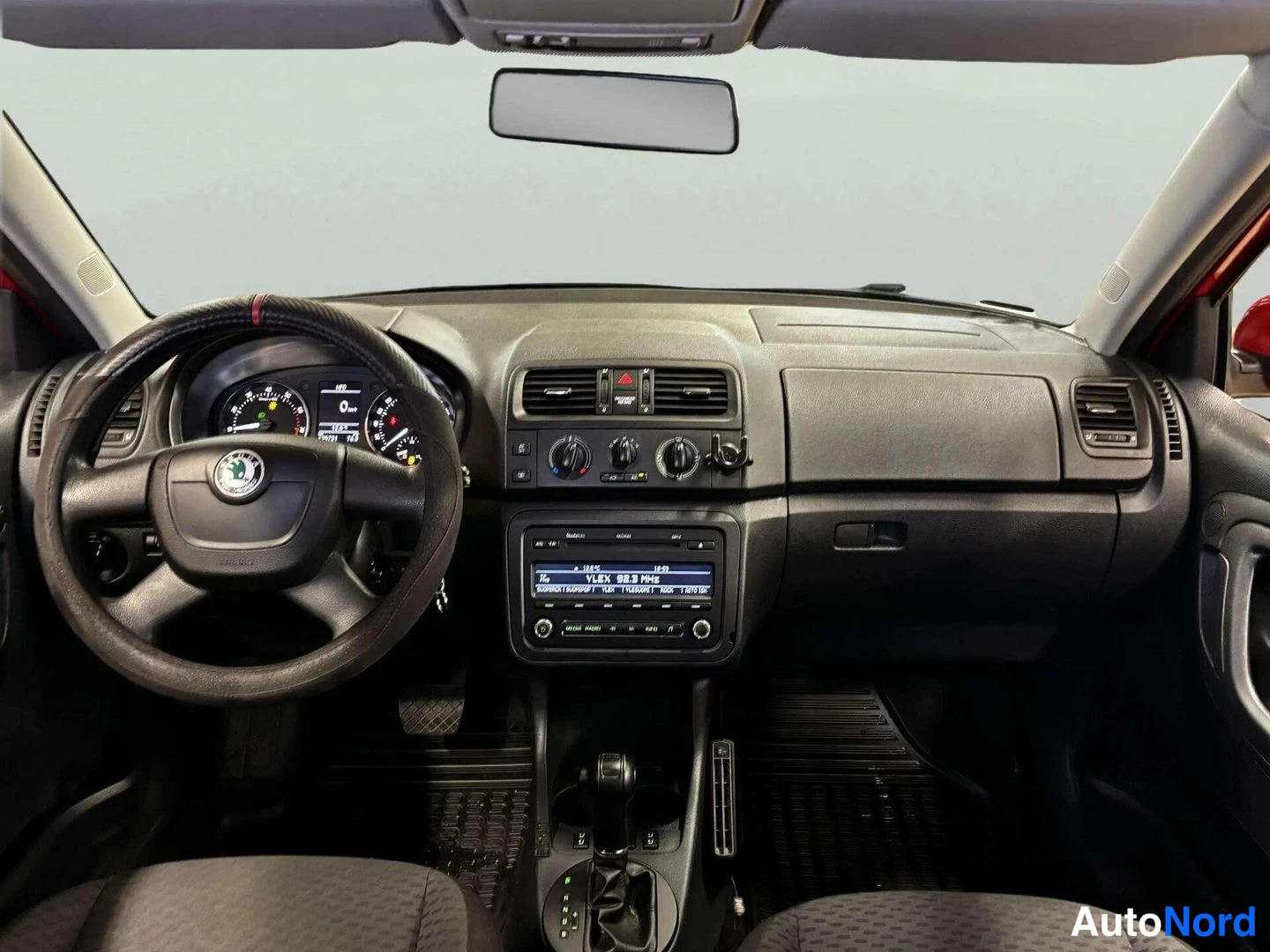 Edestä: Kojelauta ja tuulilasi suoraan edestä: ohjauspyörä Skoda-tunnuksella, mittaristo, radio-näyttö ja keskitunnelin vaihteenvalitsin näkyvät.