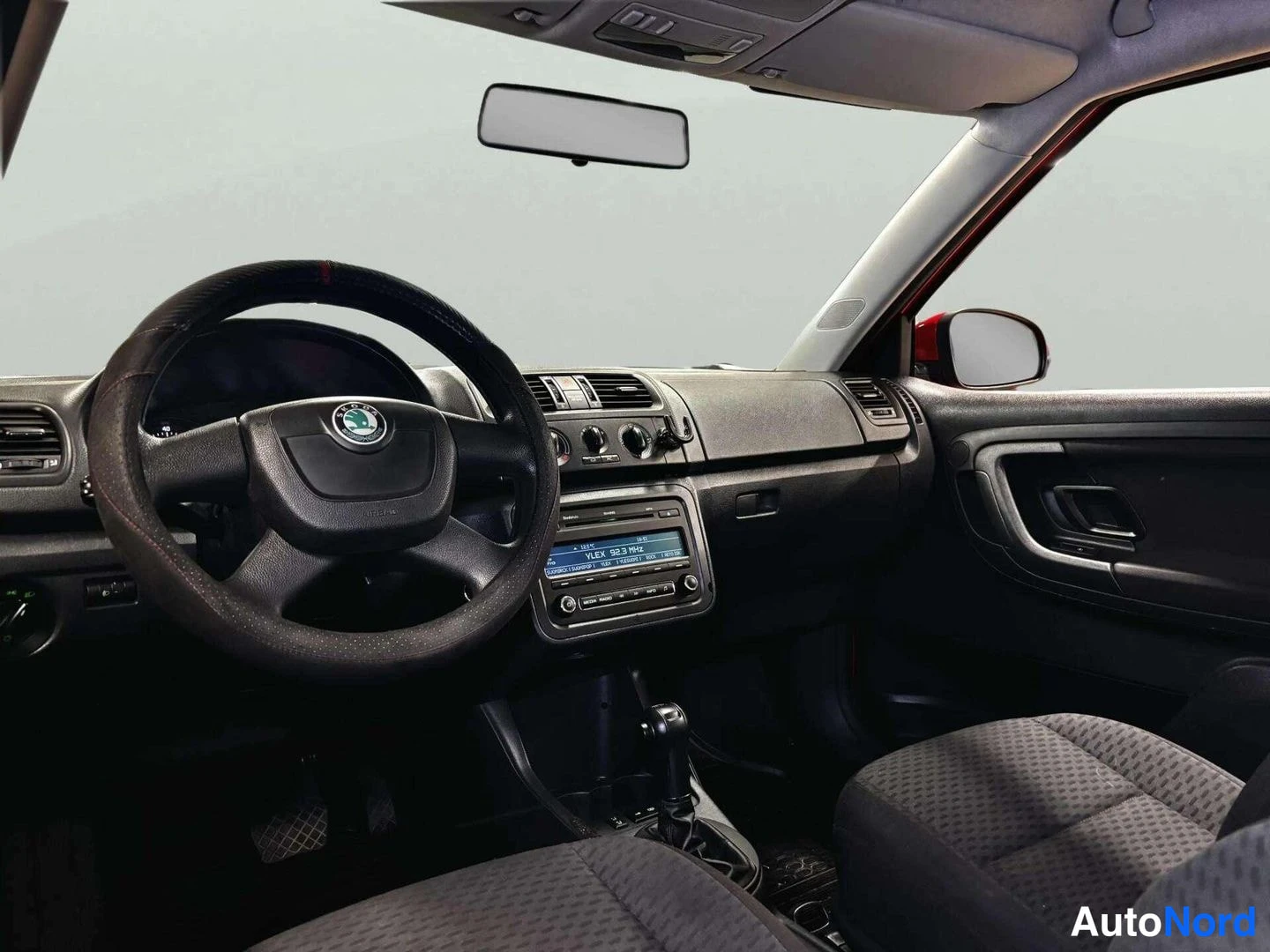 Edestä: Ohjaamo kuljettajan puolelta: Skoda-merkkinen ohjauspyörä, tumma kojelauta, radio-näyttö ja vaihteenvalitsin näkyvät; ulkopuoli ei ole kuvassa.