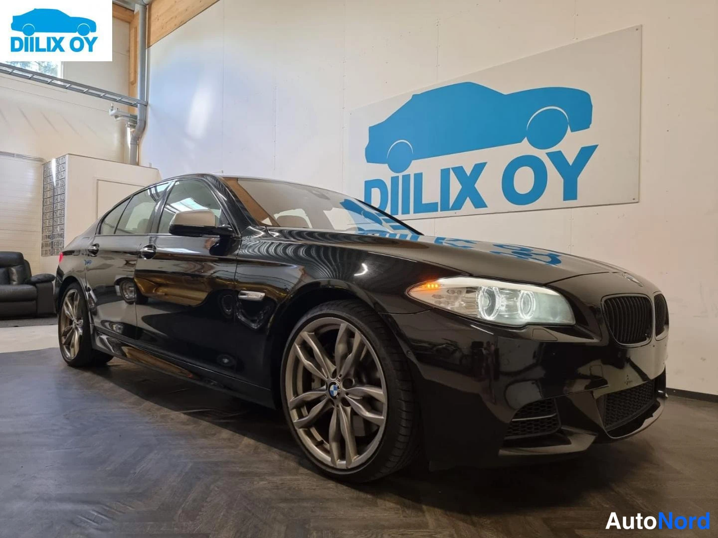 Etuviisto, nokka oikealle: Musta BMW 5-sarjan sedan edestä oikealta kuvattuna sisätiloissa, munuaisgrilli ja ajovalot näkyvissä.