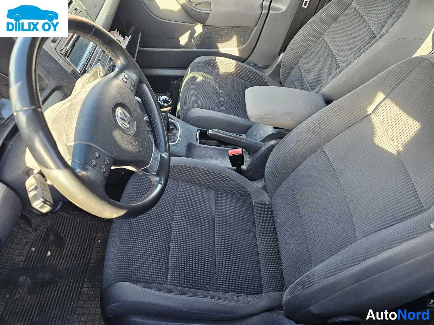 Ohjaamo: Interior cabin view showing front seats and steering wheel from passenger side angle. Dark fabric seats visible with central armrest between them. Multi-functio