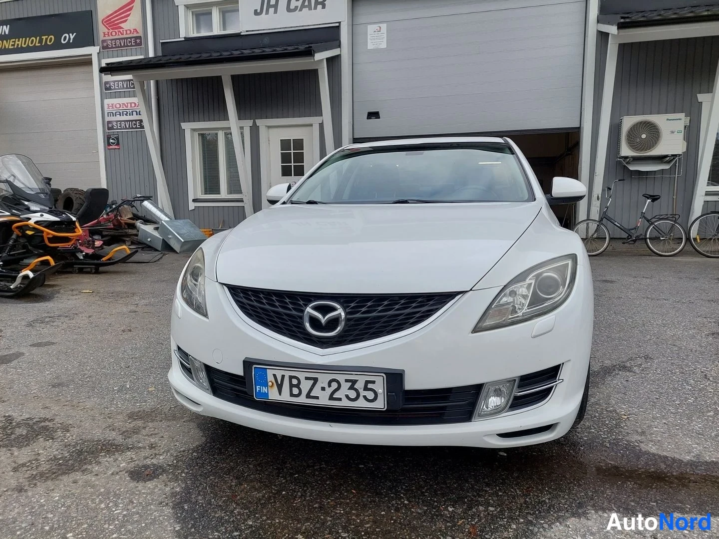 Edestä: Valkoisen Mazda 6 -sedan etupää suoraan edestä; rekisterikilpi ja Mazda-merkki etusäleikössä.
