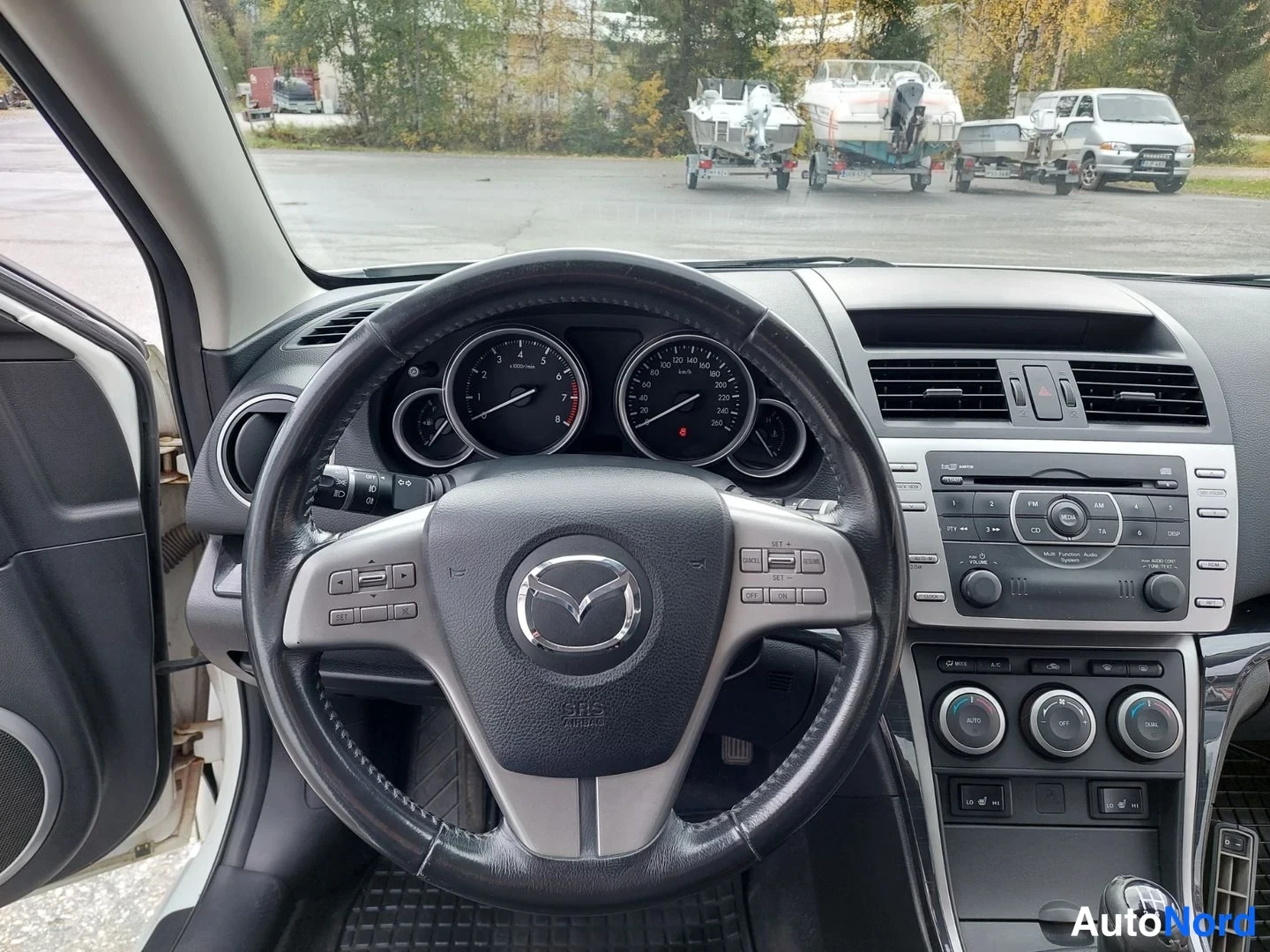 Muu: Lähikuva Mazda-ohjauspyörästä ja mittaristosta; radiopaneelissa CD-merkintä ja ilmastointisäätimet AUTO-napeilla.