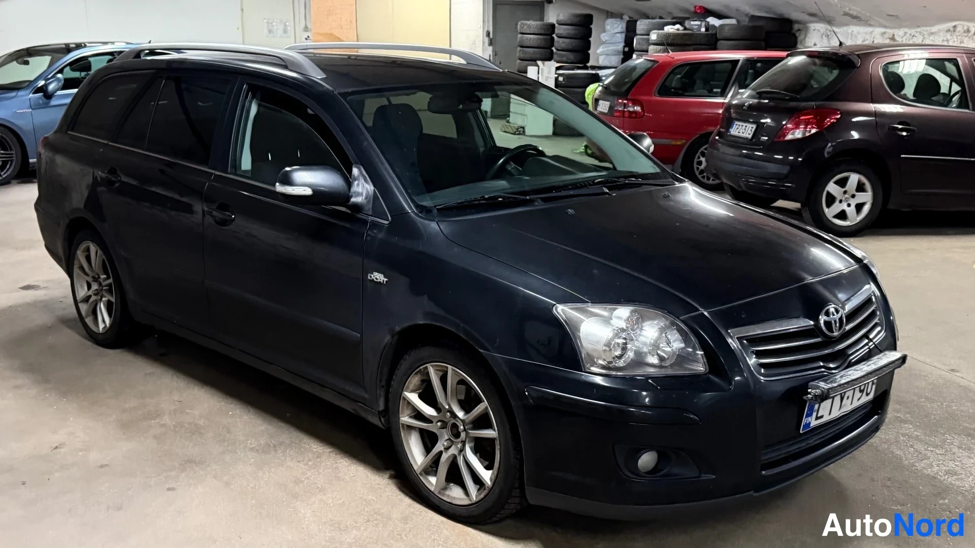 Etuviisto, nokka oikealle: Musta Toyota Avensis -farmari etukulmasta oikealta, hallissa, hopeavanteet ja kattokaiteet.