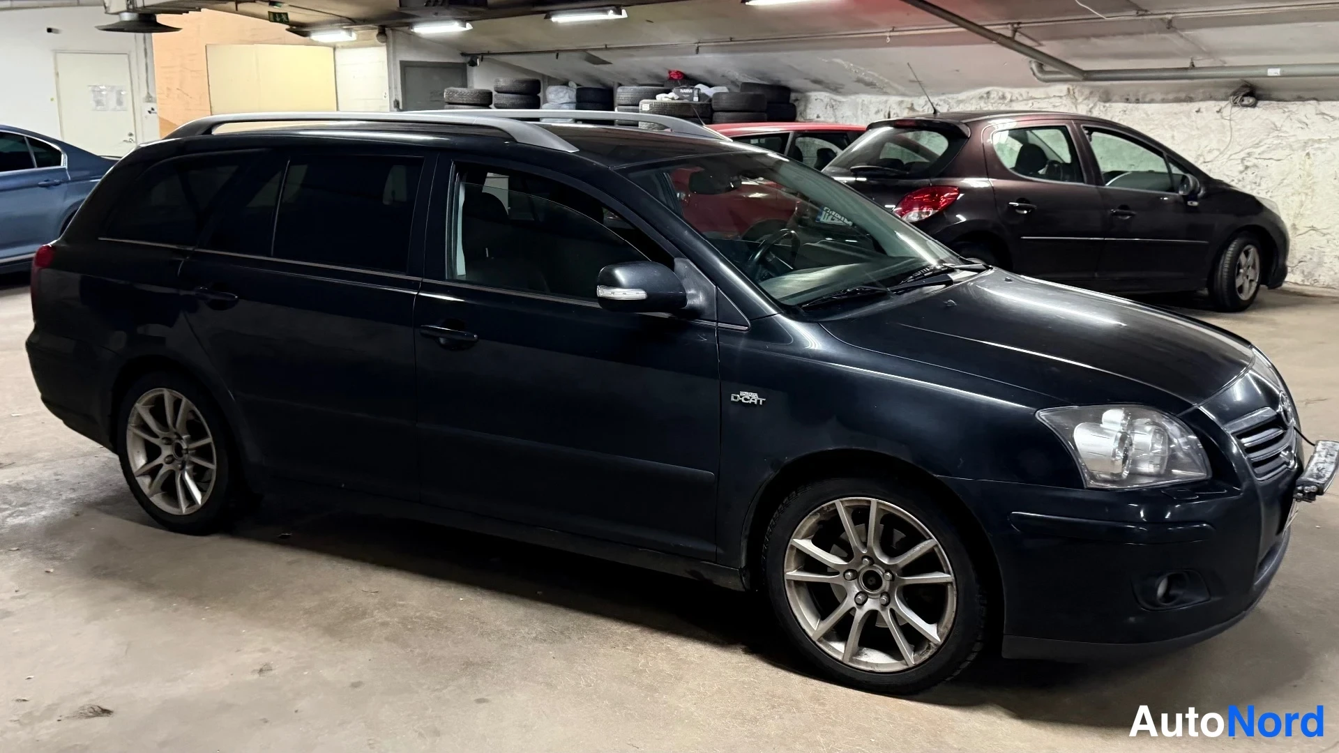 Etuviisto, nokka oikealle: Musta Toyota Avensis -farmari sivulta etukulmaan, halli, kattokaiteet, hopeavanteet.