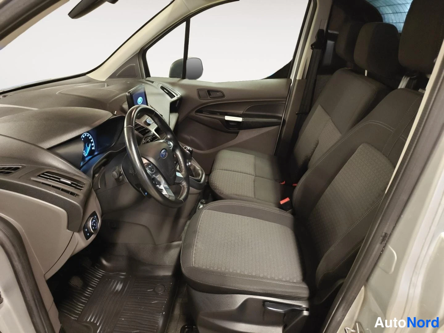 Muu: Ford Transit Connectin ohjaamo sivulta: ohjauspyörä, vaihdin, tummat kankaat istuimet ja kuormatilaan avautuva aukeama.