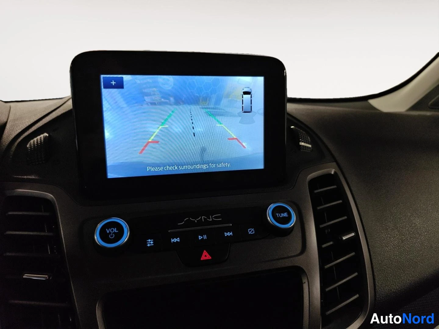 Muu: Ford Transit Connectin kojelaudan SYNC-näyttö näyttää peruutuskameran kuvaa ohjausviivoineen ja tekstillä 'Please check surroundings for safety.'