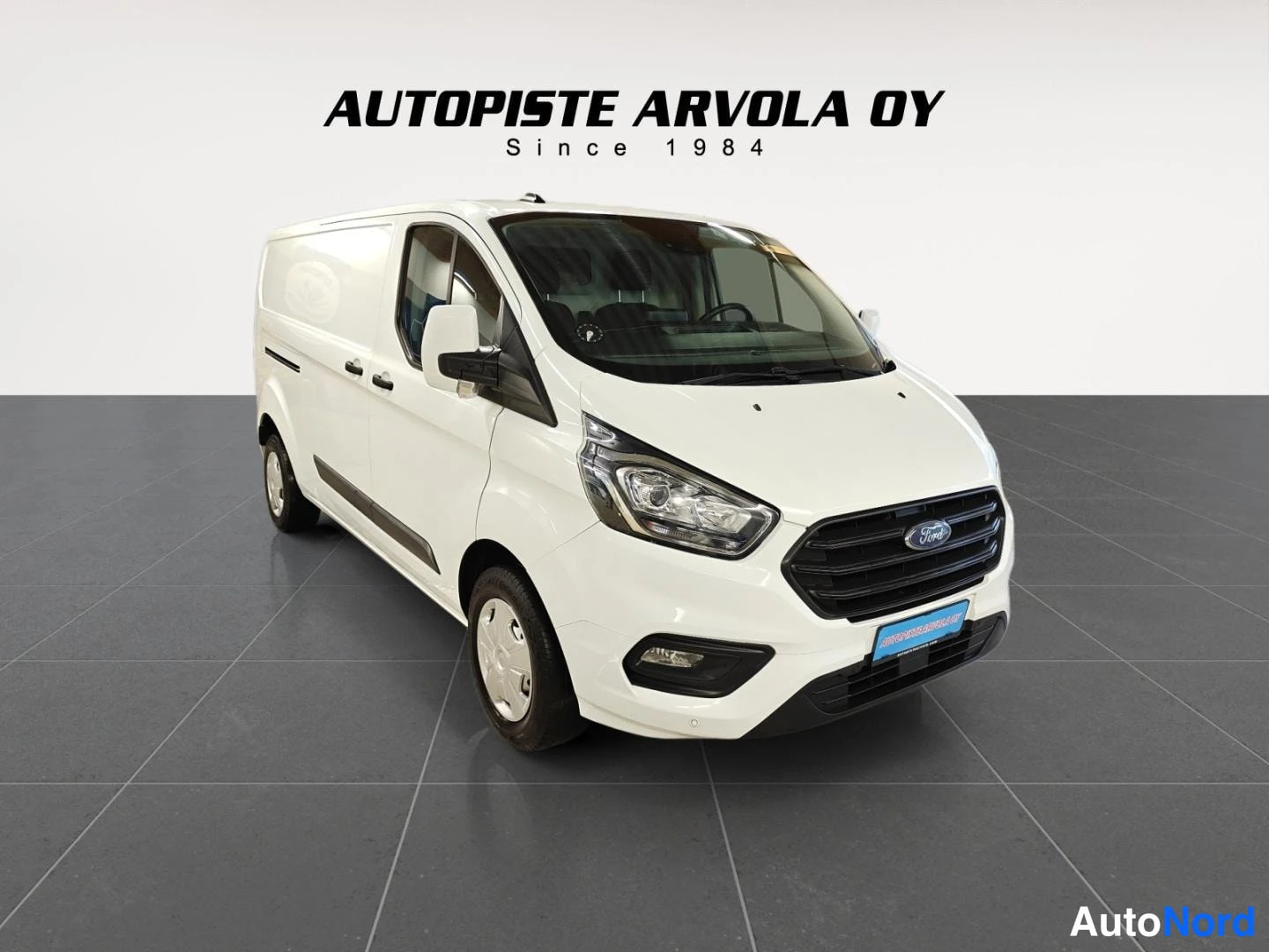 Etuviisto, nokka oikealle: Valkoinen Ford Transit Custom -pakettiauto kolmivartalokuvassa edestä oikealta, etusäleikkö ja ajovalot näkyvissä.