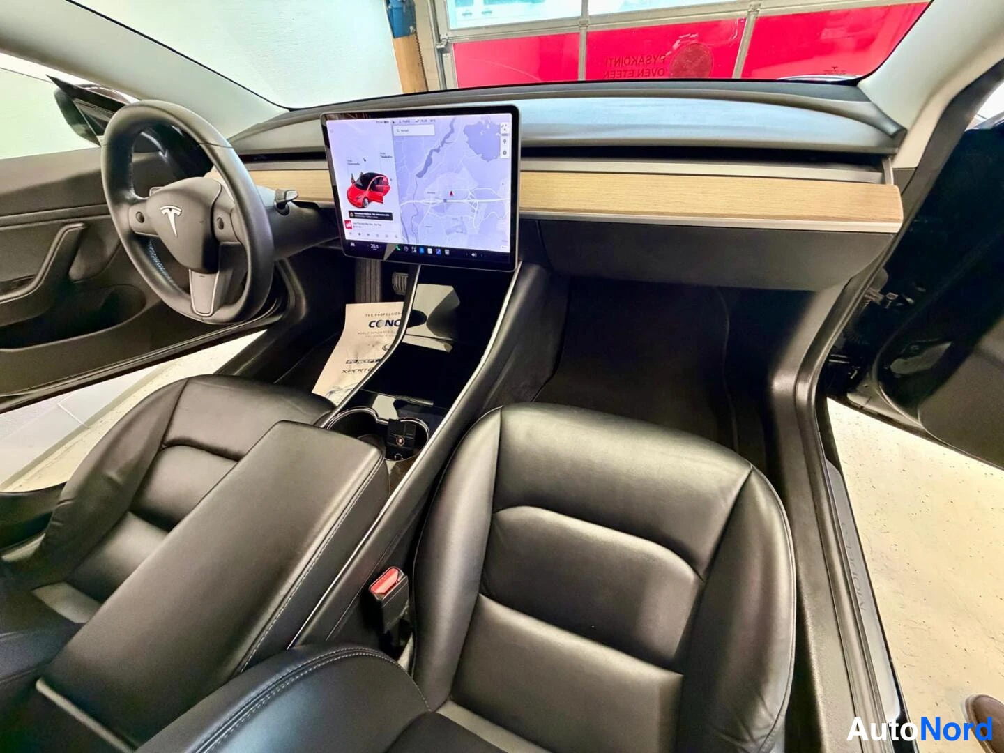 Muu: Kohtisuora näkymä Tesla Model 3:n kojelautaan: ohjauspyörä, keskeinen karttaa näyttävä 15 tuuman näyttö, vaalea puukoriste ja mustat penkit.