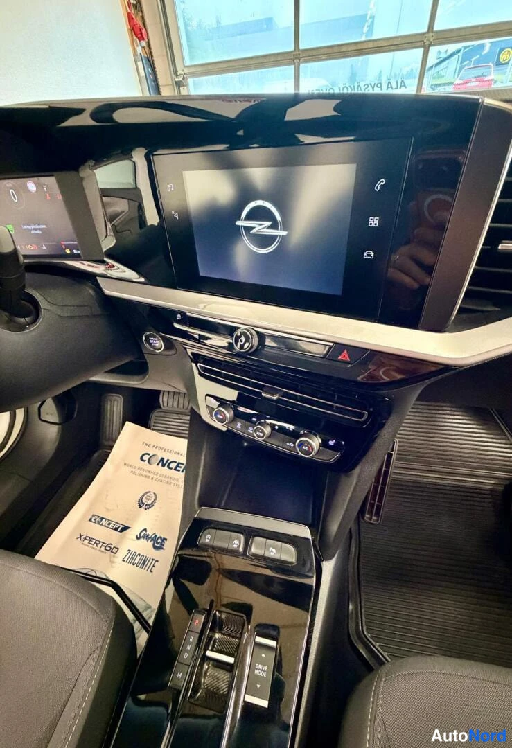 Muu: Keskikonsolin infotainment-näyttö Opel-logolla käynnissä, alle ilmastointisäätimet, dokumentti lattialla ja automaattivaihteiston valitsin.