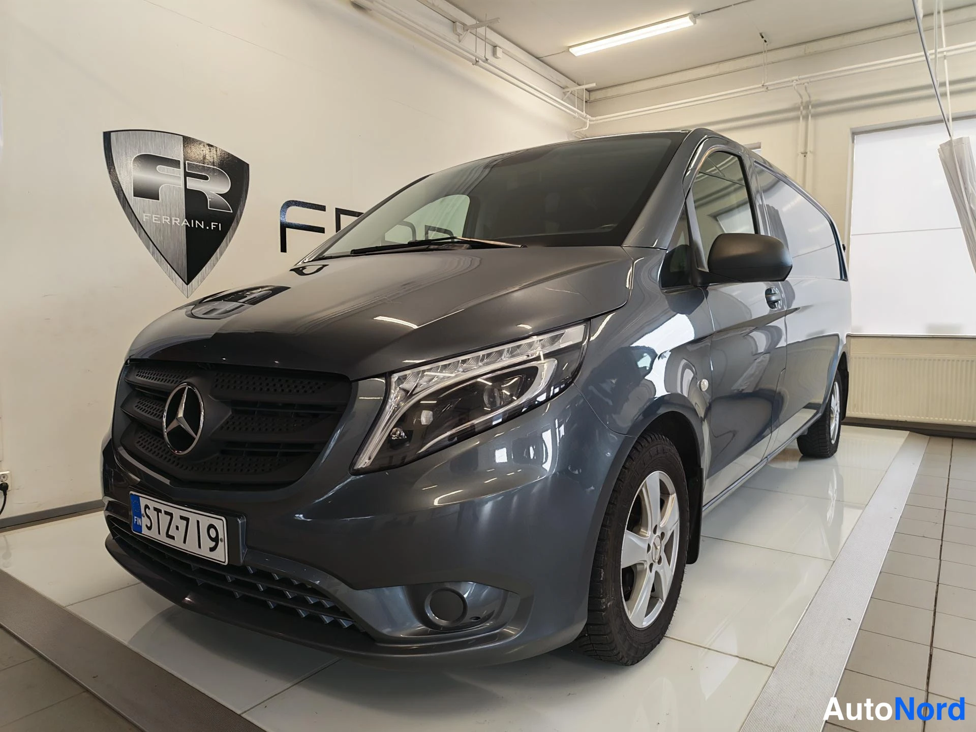 Etuviisto, nokka vasemmalle: Mercedes-Benz Vito -pakettiauto tummanharmaana edestä oikealta kuvattuna sisätiloissa, rekisterikilpi.