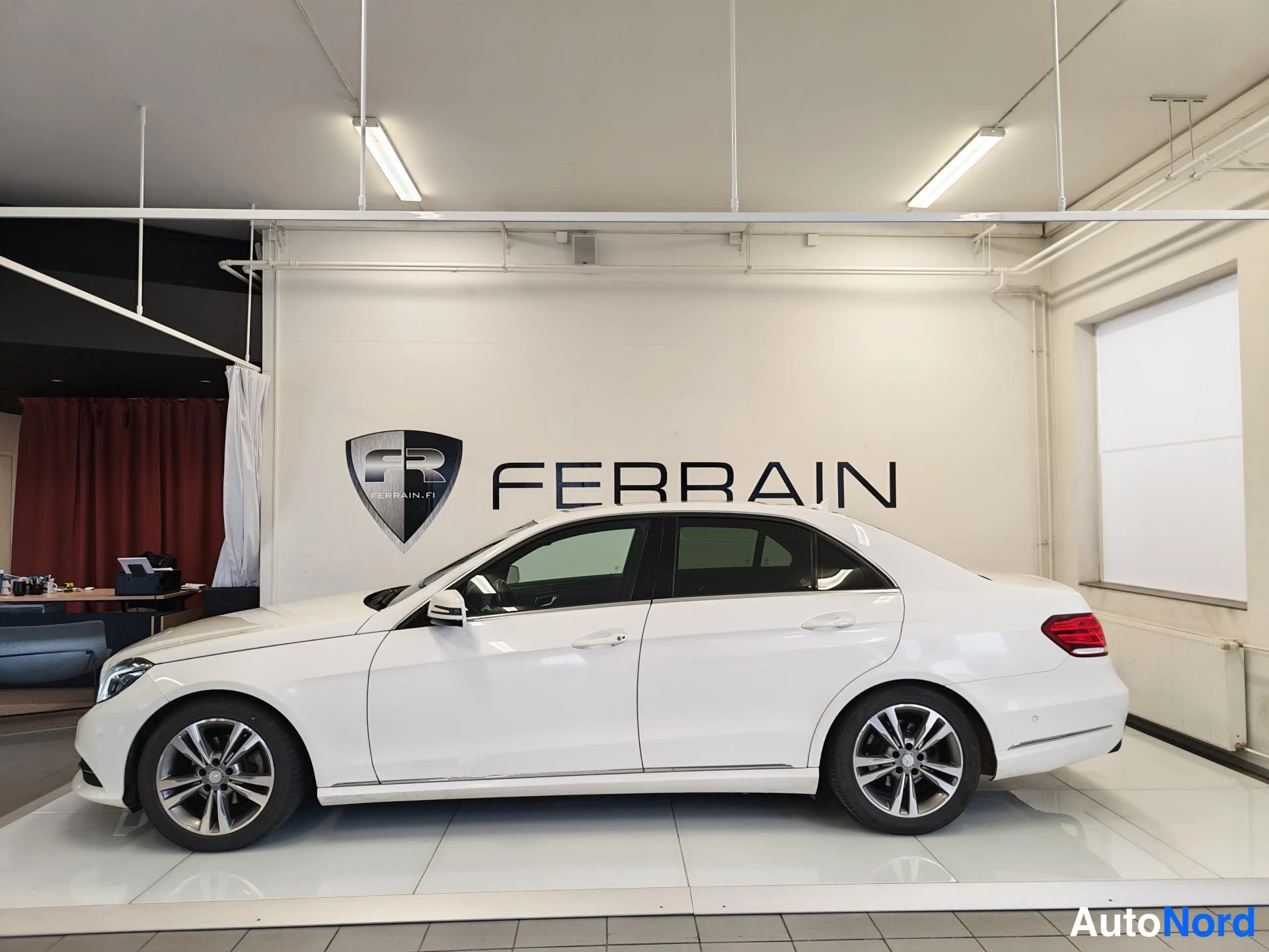 Sivulta: Valkoinen Mercedes-Benz E-sarjan sedan sivusta oikealta kuvattuna näyttelytilassa, neljä ovea ja kevytmetallivanteet näkyvissä.