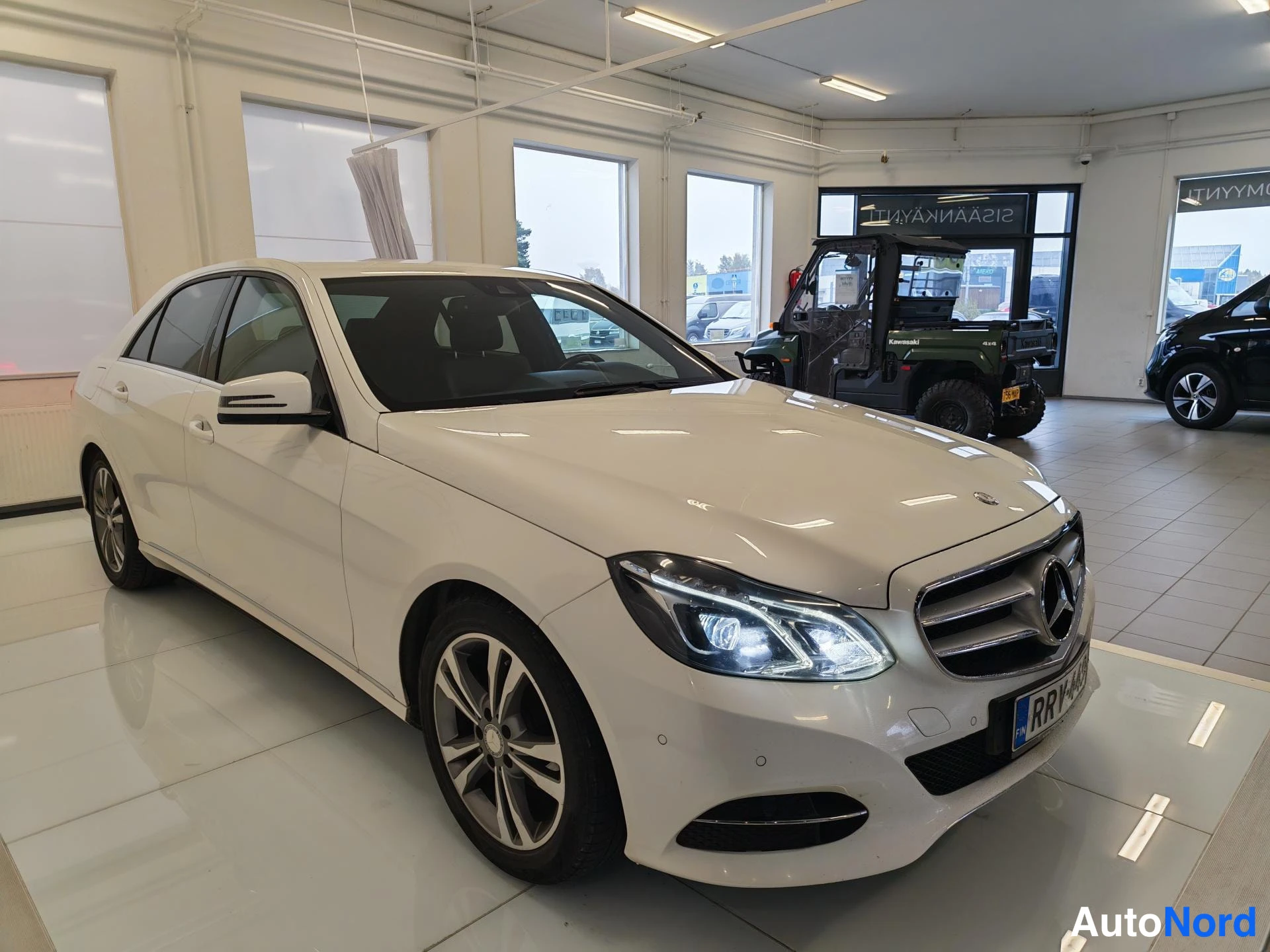 Etuviisto, nokka oikealle: Auton etukulma sivulta: valkoinen Mercedes-Benz -sedan neljällä ovella, keula oikealla, ajovalot päällä ja rekisterikilpi RR-44.