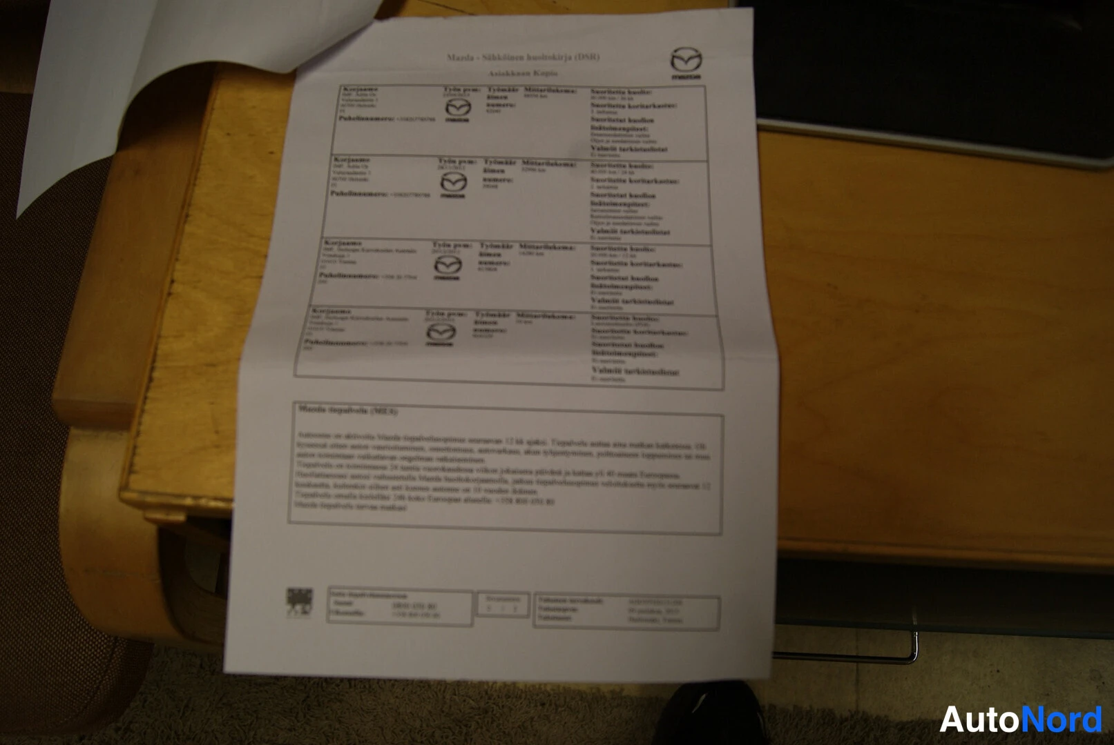 Muu: Tulostettu Mazda – sähköinen huoltokirja (DSR) -asiakirja, jossa huoltotietueita ja Huolto tapahtuivat (Mikkeli) -osio, paperi puupöydällä.