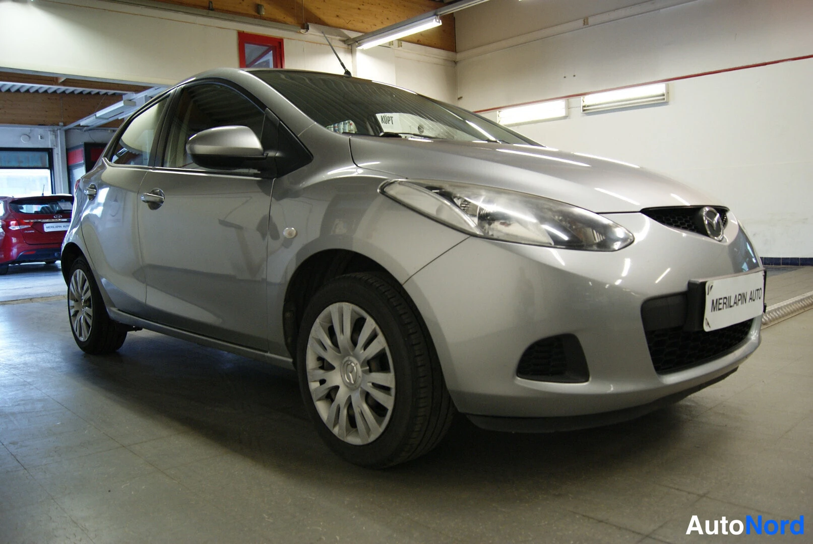 Etuviisto, nokka oikealle: Hopeanharmaa Mazda 2 autotilassa, keula oikealla ja kylki näkyvissä.