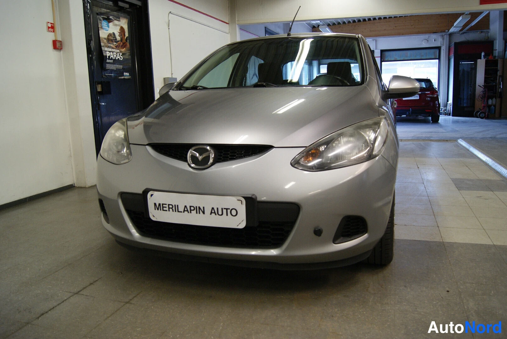 Edestä: Hopeanharmaa Mazda 2 etupuskureineen, etugriffineen ja ajovaloineen sisätiloissa.