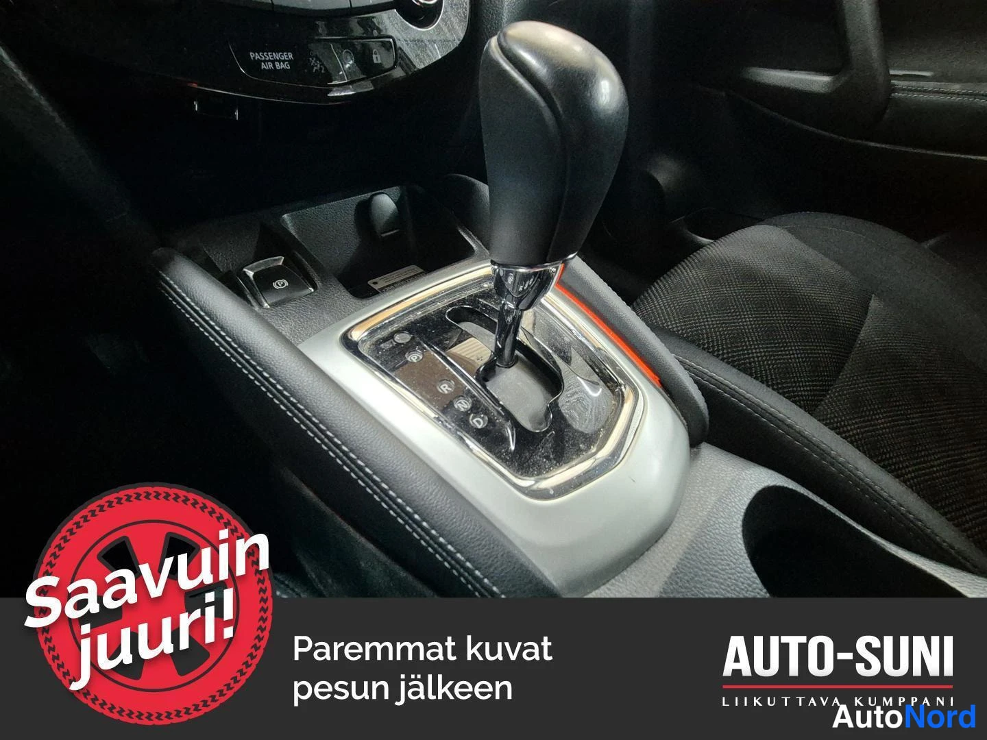Muu: Auton keskikonsolin automaattivaihteiston vaihdevipu ja ympärillä olevia painikkeita, taustalla tummaa istuinta; kuvassa myös AUTO-SUNI-teksti.