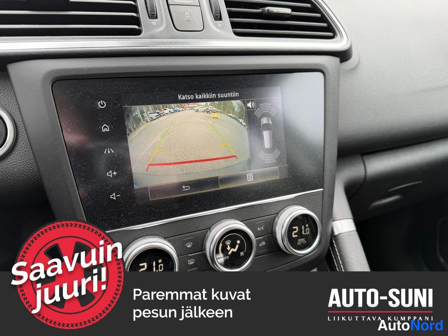 Muu: Kojelaudan infotainment-näyttö, jossa näkyy peruutuskameran kuva tekstillä Katso kaikkiin suuntiin sekä automaattipaisteasettimen pyörät.