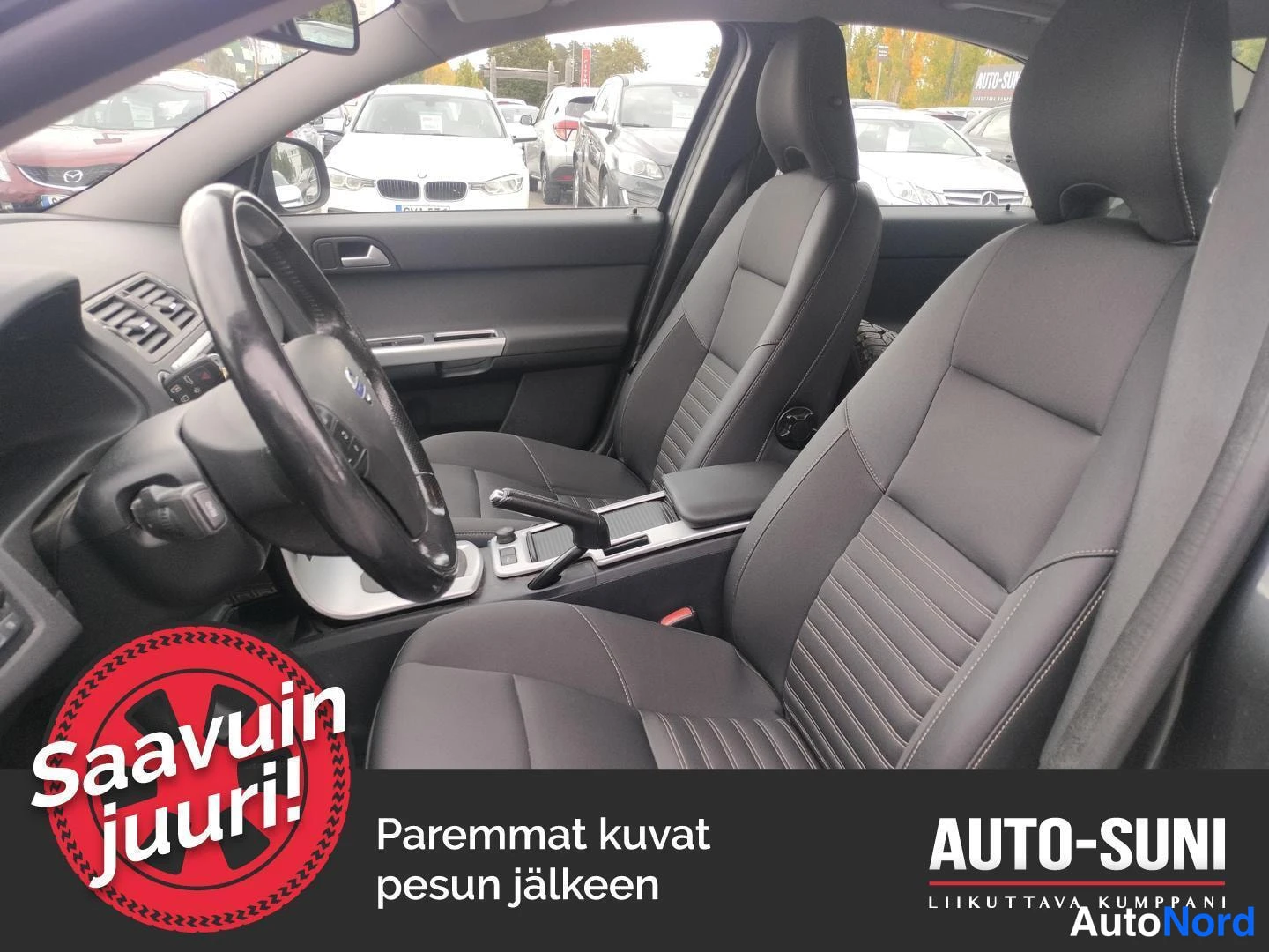 Muu: Sisätila kuljettajan puolelta: kaksi etuistuinta, ohjauspyörä, käsijarru ja vaihdekeksi keskikonsolissa, taustalla näkyy autojäonoja.