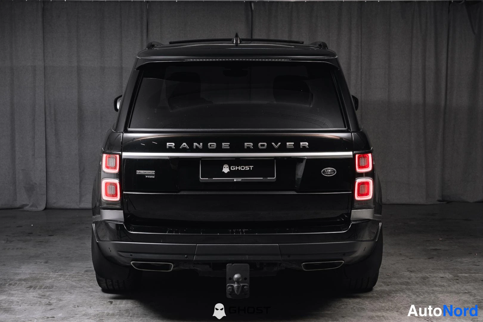 Takaa: Mustan Range Rover -maasturin suora takanäkymä, RANGEROVER-teksti takaluukussa, punaiset takavalot ja perävakokuppi harmaata verhoselia vasten.
