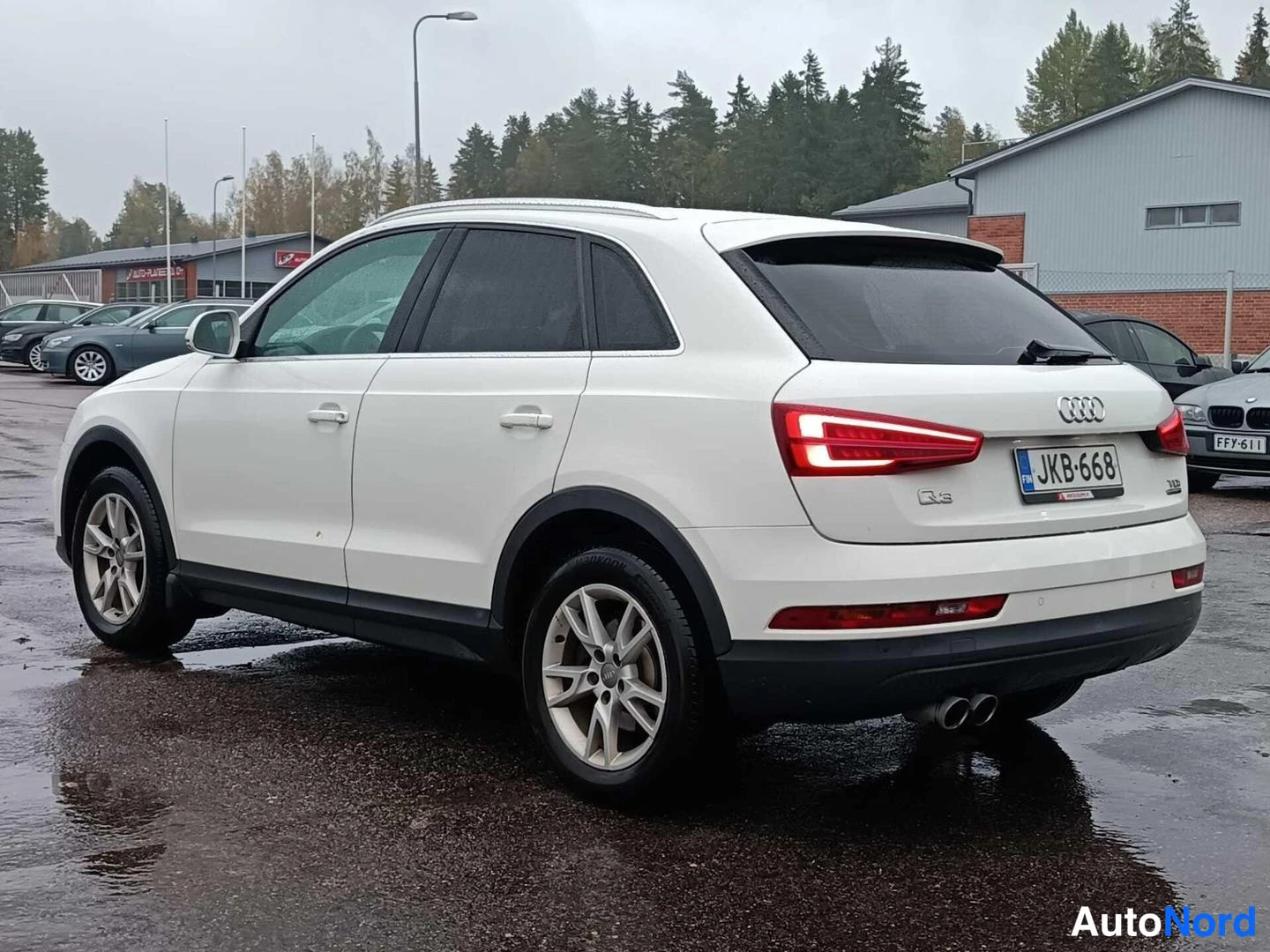 Takaviisto: Valkoisen Audin takaosan ja vasemman sivun kolmasosan kuva märällä parkkipaikalla; rekisterikilpi.