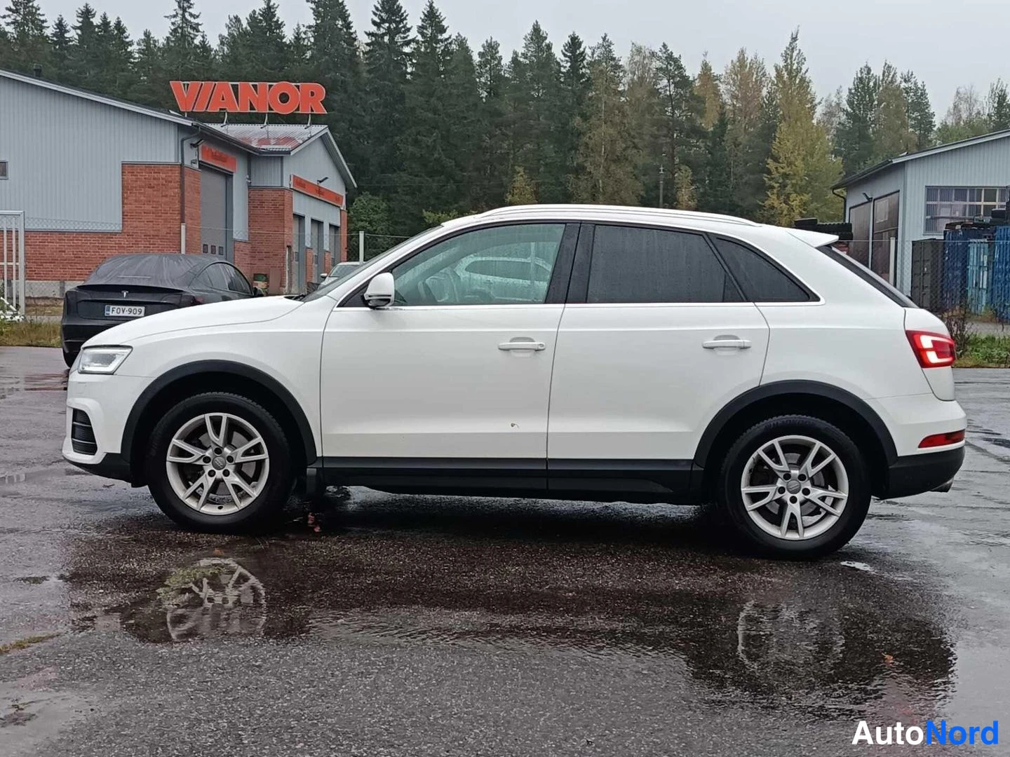 Sivulta: Valkoinen Audi sivusta etukolmasosasta, keula kuvan vasemmassa reunassa; sateinen asfalttipiha ja Vianor-rakennus taustalla.