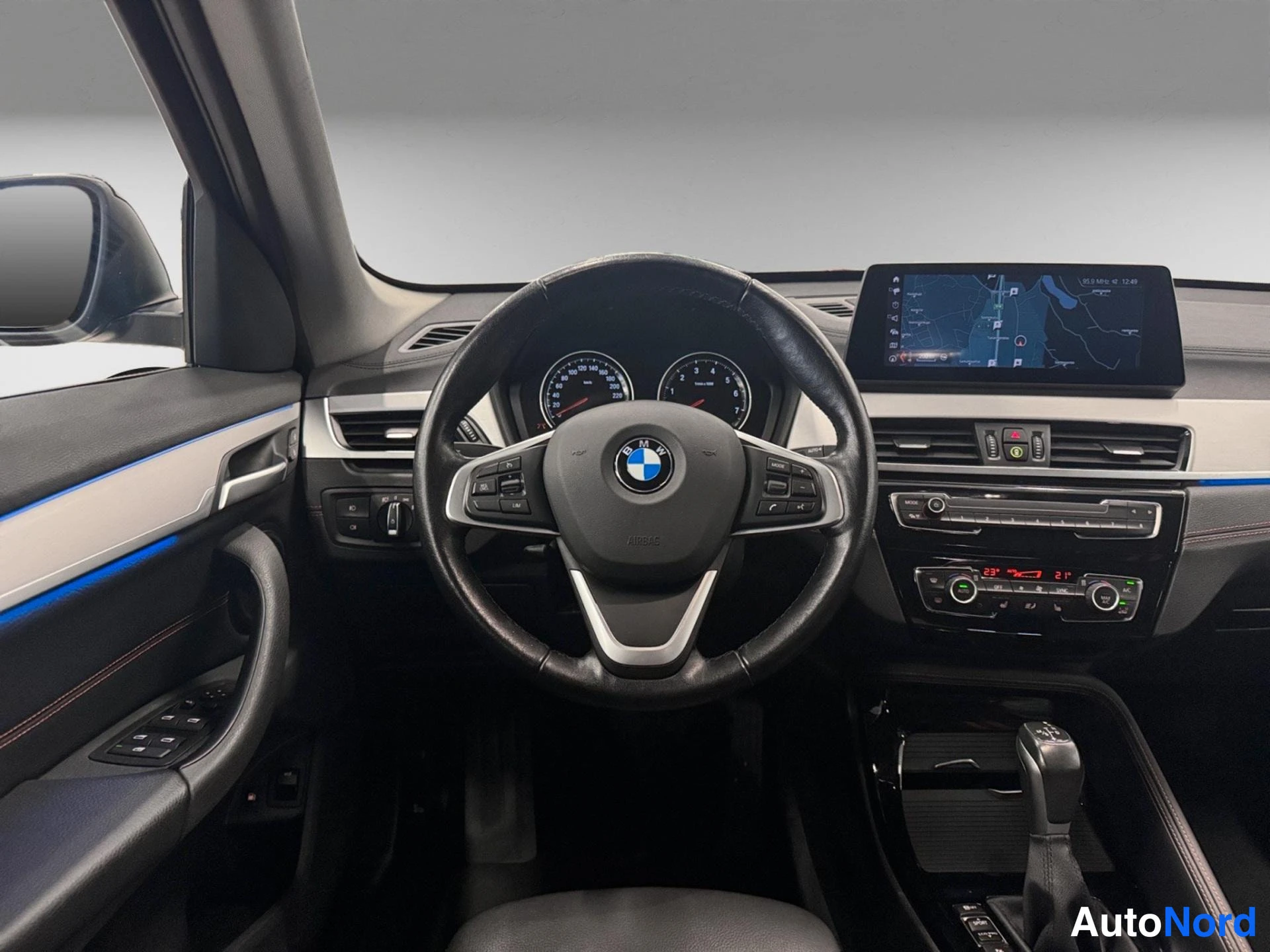 Edestä: Henkilöauton ohjaamo: BMW-tunnuksella varustettu ohjauspyörä, analogiset mittarit, karttaa näyttävä keskinäyttö, automaattisen ilmastoinnin paneeli ja automaatt