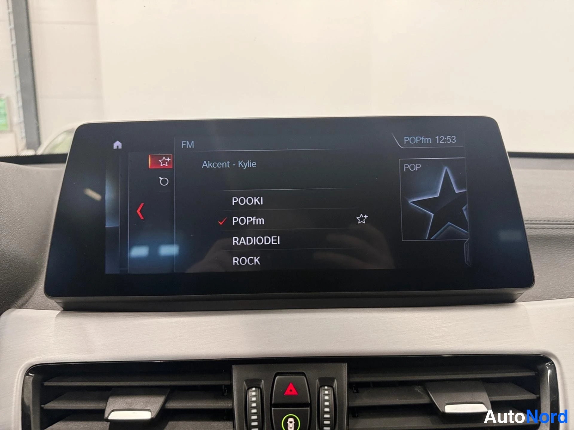 Yksityiskohta: Infotainment-näyttö kojelaudassa näyttää FM-radiolistan, jolla asemat POOKI, POPfm, RADIODEI ja ROCK; valittuna POPfm, soiva kappale 'Akcent - Kylie'.