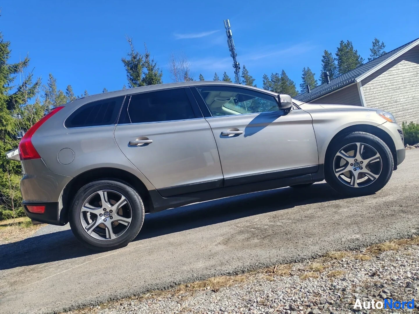 Takaviisto: Volvo XC60 -farmariauto sivulta-takaa kuvattuna hiekkaisella pihalla, keltainen takavalo ja viisipaisteiset vanteet näkyvät.